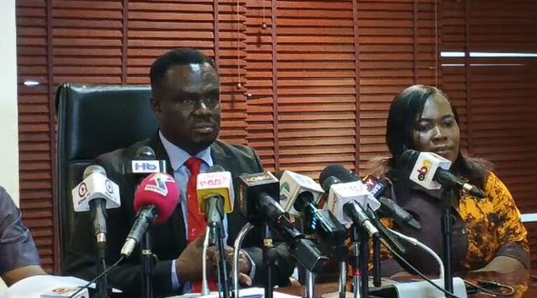 Man in a suit and red tie and a woman in a patterned top sit at a table during a press conference, microphones in front of them, wooden blinds in the background.