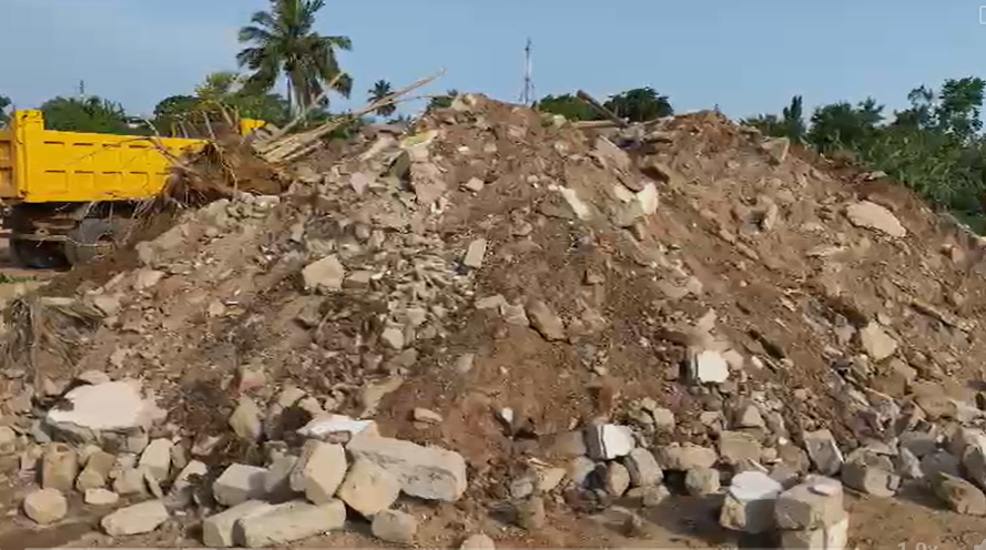 Large mound of dirt and rubble at a construction site, with a yellow dump truck on the left and palm trees in the background.