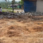 Open dirt lot with piles of soil and scattered rubble, modest houses in the background, and a person standing near the debris.