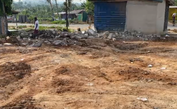 Open dirt lot with piles of soil and scattered rubble, modest houses in the background, and a person standing near the debris.