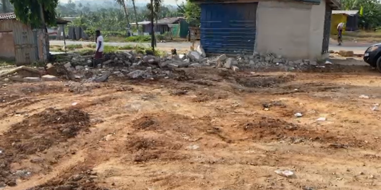 Open dirt lot with piles of soil and scattered rubble, modest houses in the background, and a person standing near the debris.
