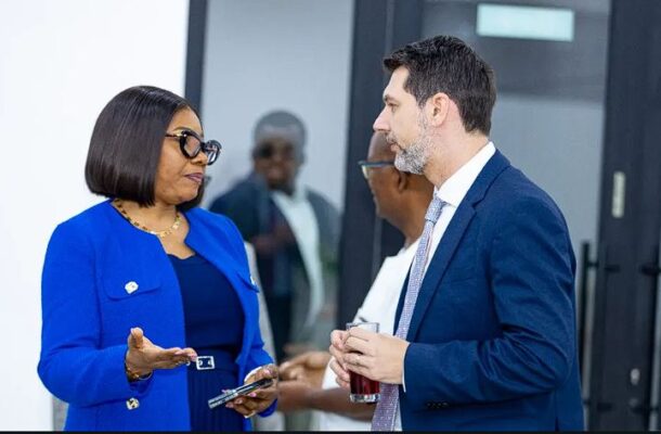 Two professionals in a blue blazer and a dark suit converse in a modern office lobby, one holding a phone and the other a drink.