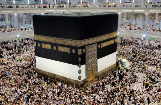 The Kaaba cubical black building surrounded by thousands of pilgrims inside the Grand Mosque in Mecca, with gold band and door visible.