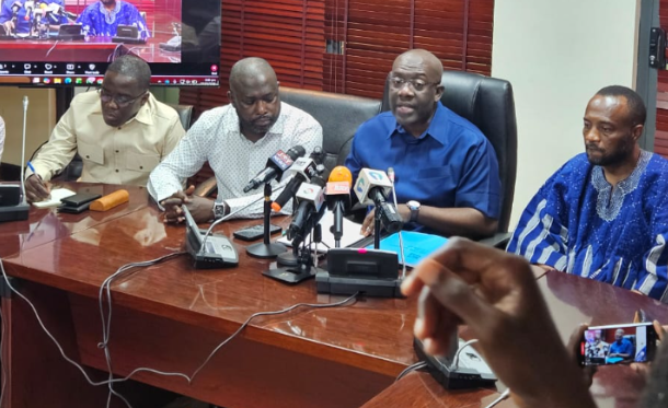 Four men seated at a wooden table during a press conference with multiple microphones in front of them.