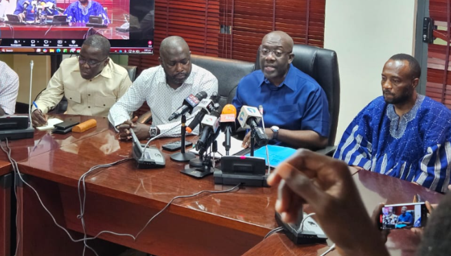 Four men seated at a wooden table during a press conference with multiple microphones in front of them.