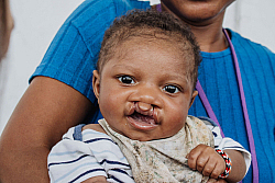 Smiling baby being held by an adult wearing a blue shirt.