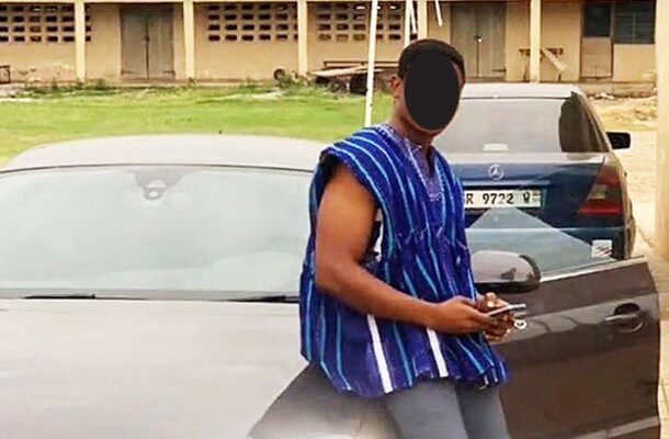 Man in a blue striped sleeveless shirt leaning against a silver car in a parking lot, looking at his phone; his face is obscured.