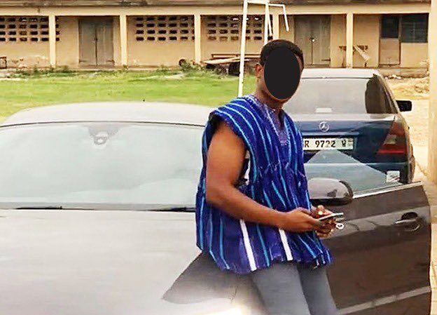 Man in a blue striped sleeveless shirt leaning against a silver car in a parking lot, looking at his phone; his face is obscured.