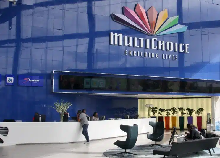 Reception area of a MultiChoice building with a blue exterior and colorful logo, staff behind the counter and visitors seated nearby