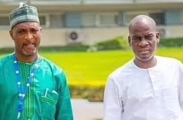Two men stand outdoors, one in a green traditional outfit with a cap and the other in a white tunic, both looking toward the camera.
