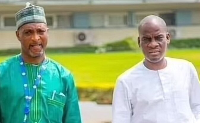 Two men stand outdoors, one in a green traditional outfit with a cap and the other in a white tunic, both looking toward the camera.
