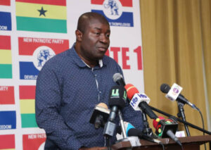 Man speaking at a podium with multiple microphones during a press conference, backdrop shows party banners in red, yellow, and blue.