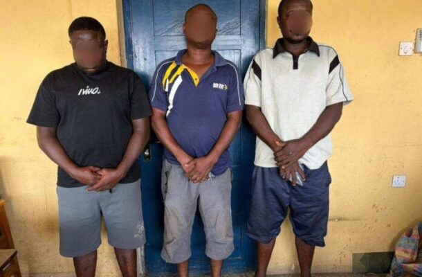 Three men with blurred faces stand side by side against a yellow wall and blue door, hands clasped in front.