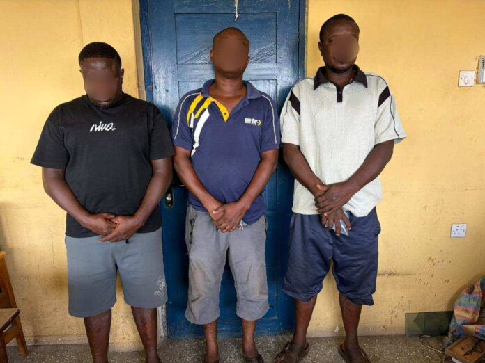 Three men with blurred faces stand side by side against a yellow wall and blue door, hands clasped in front.