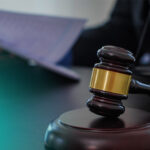 Close-up of a wooden judge's gavel with a gold band resting on a sound block on a desk, blurred courtroom background.