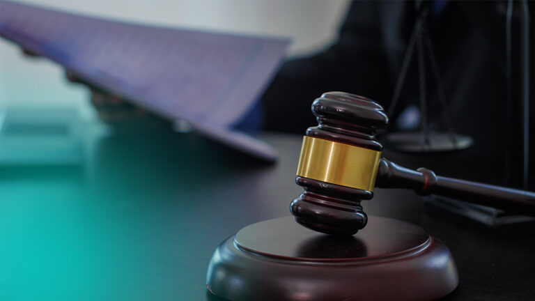 Close-up of a wooden judge's gavel with a gold band resting on a sound block on a desk, blurred courtroom background.