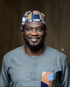 Portrait of a smiling man wearing a patterned cap and a gray traditional shirt with a geometric pocket, against a dark blurred background.