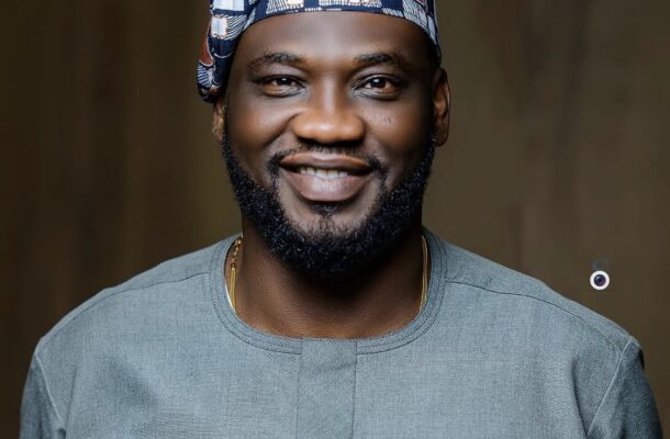 Portrait of a smiling man wearing a patterned cap and a gray traditional shirt with a geometric pocket, against a dark blurred background.