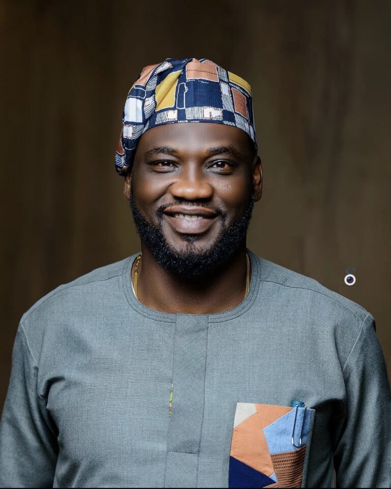 Portrait of a smiling man wearing a patterned cap and a gray traditional shirt with a geometric pocket, against a dark blurred background.