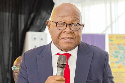 Man in a navy suit and red tie speaks into a handheld microphone at an indoor event.
