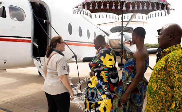 Staff member helps travelers in colorful African attire board a small white private jet under a decorative parasol on the tarmac.