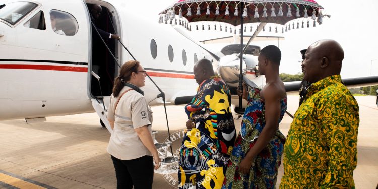 Staff member helps travelers in colorful African attire board a small white private jet under a decorative parasol on the tarmac.