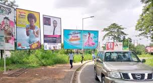 Colorful roadside billboards with models and ads along a curved street; pedestrians walking and a silver SUV nearby.