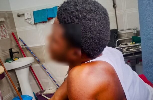Back view of a shirtless person with short curly hair resting in a bathroom, facing away from the camera, with a sink and cleaning supplies in the background.