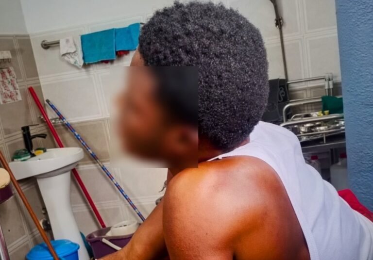 Back view of a shirtless person with short curly hair resting in a bathroom, facing away from the camera, with a sink and cleaning supplies in the background.
