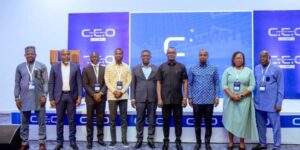 Ten professionals in business attire stand in a row on a conference stage with a blue branded backdrop and lanyards.4