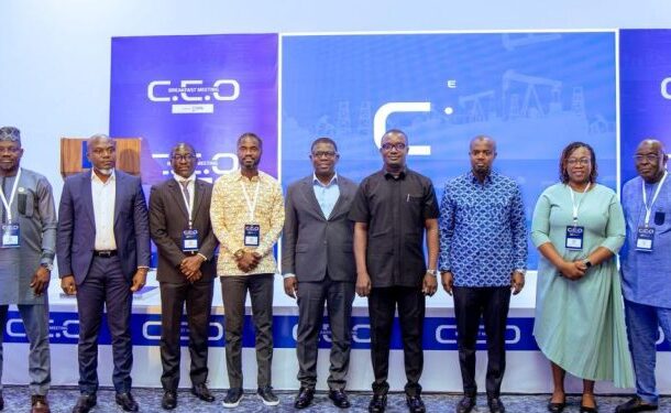 Ten professionals in business attire stand in a row on a conference stage with a blue branded backdrop and lanyards.4