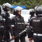 Ghana Police riot squad officers in protective gear standing on a street, one in the center holding a baton.