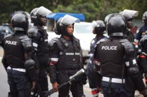 Ghana Police riot squad officers in protective gear standing on a street, one in the center holding a baton.