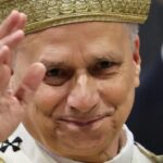 Ceremonial figure in a white robe and gold crown, smiling and saluting with his right hand.