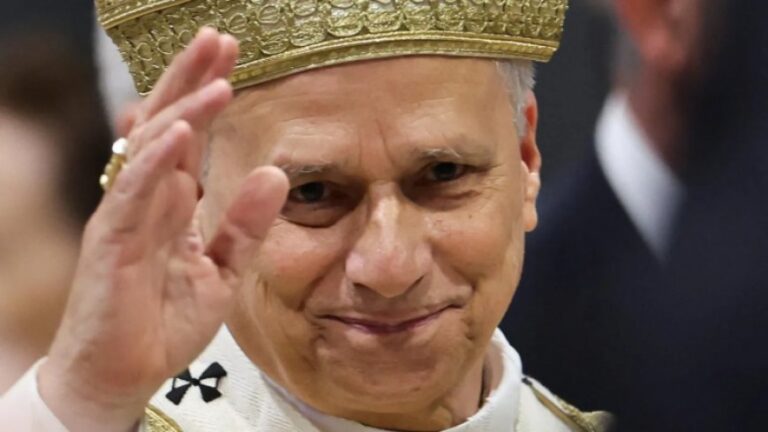 Ceremonial figure in a white robe and gold crown, smiling and saluting with his right hand.
