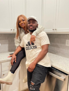 Couple posing in a bright kitchen: woman sits on the counter behind man with an arm around his shoulder; both smile, man in a beige Adidas shirt and cap.