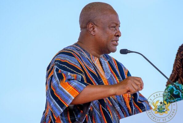 Man in colorful traditional fabric speaks into a microphone at a podium, with the Republic of Ghana seal visible on the desk.