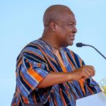 Man wearing colorful striped kente cloth speaks at a podium with a microphone, against a light blue background.