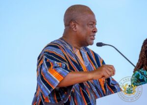 Man wearing colorful striped kente cloth speaks at a podium with a microphone, against a light blue background.