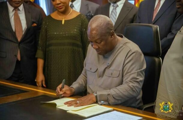 Man in a gray shirt signs a document at a desk as others in formal attire look on; official seal visible in bottom corner.