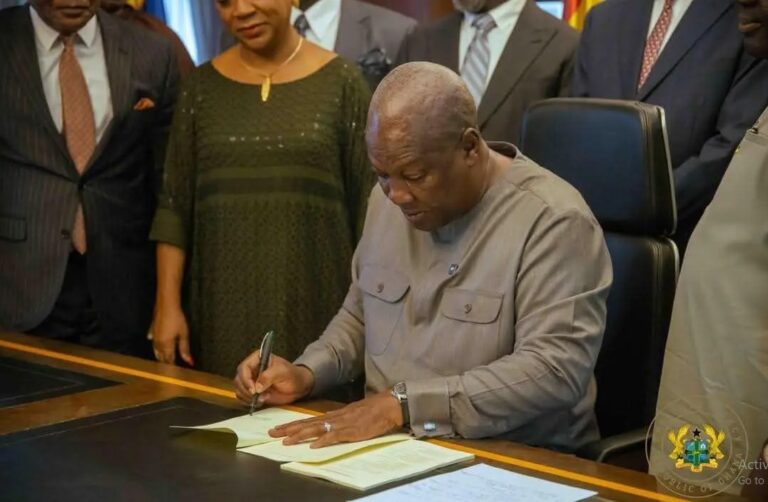 Man in a gray shirt signs a document at a desk as others in formal attire look on; official seal visible in bottom corner.