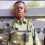 Soldier in camouflage uniform wearing red over-ear headphones and a chest radio, standing behind a wooden fence outdoors.