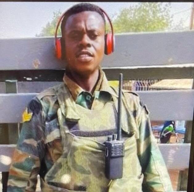 Soldier in camouflage uniform wearing red over-ear headphones and a chest radio, standing behind a wooden fence outdoors.