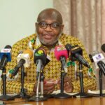 Man in a patterned yellow shirt speaks at a press conference with multiple microphones in front of him.