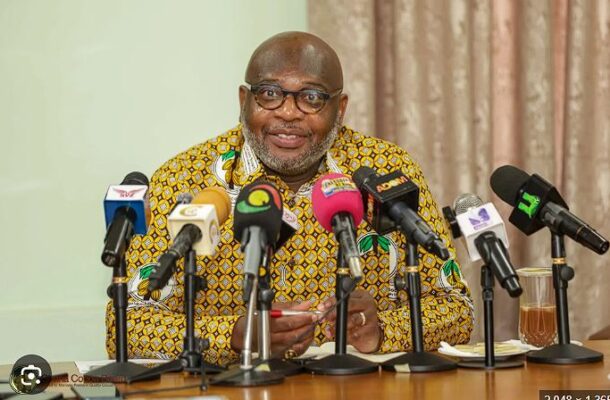 Man in a patterned yellow shirt speaks at a press conference with multiple microphones in front of him.