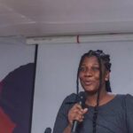 Black woman on stage speaking into a handheld microphone at a conference, with a banner in the background reading 'I Believe Global' (partially visible).