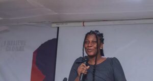Black woman on stage speaking into a handheld microphone at a conference, with a banner in the background reading 'I Believe Global' (partially visible).