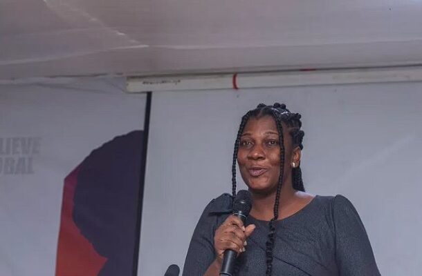 Black woman on stage speaking into a handheld microphone at a conference, with a banner in the background reading 'I Believe Global' (partially visible).