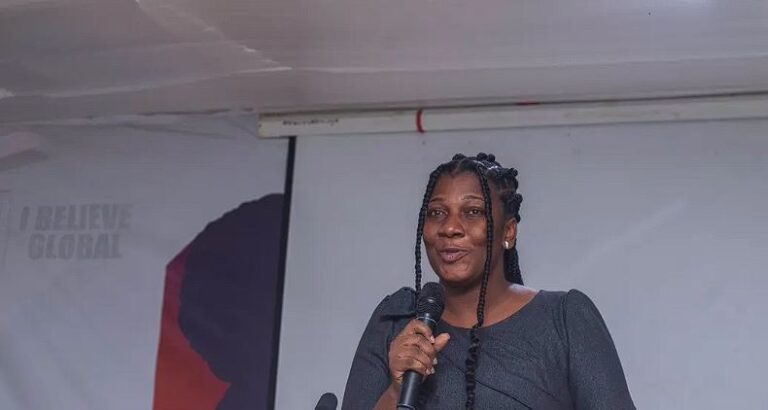 Black woman on stage speaking into a handheld microphone at a conference, with a banner in the background reading 'I Believe Global' (partially visible).