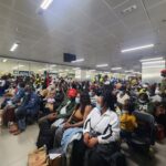 Crowd of travelers wearing masks seated in an airport waiting area with luggage, security personnel visible near counters in the background.